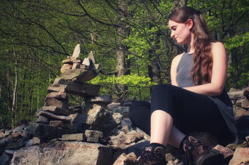Low angle view of young woman looking at stacked rocks while sitting in forest