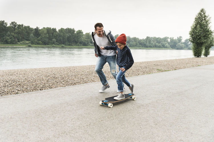 Happy father running next to son on skateboard at the riverside
