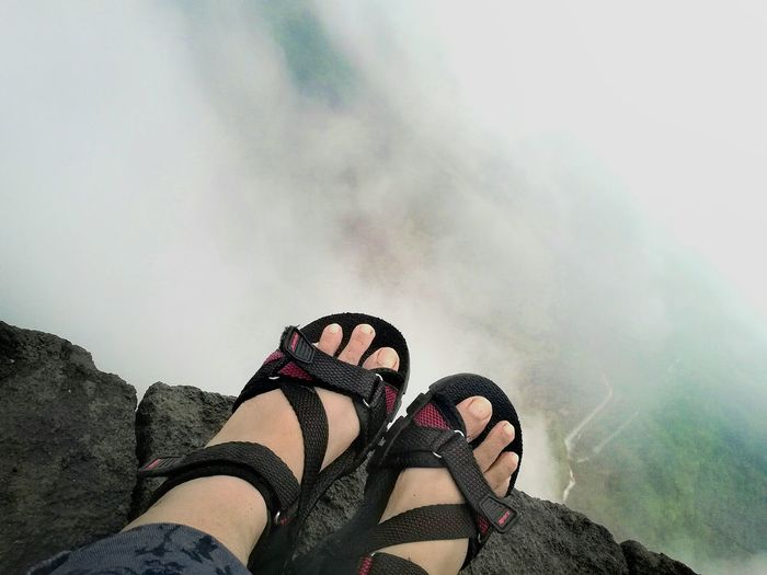 Low section of person wearing scandals on cliff amidst clouds