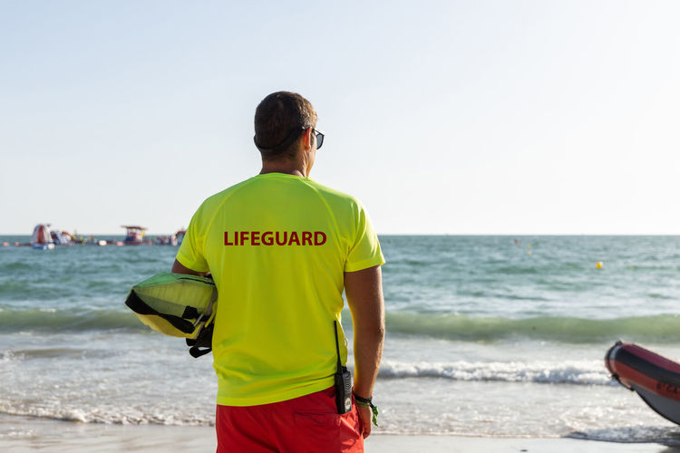 Back view of anonymous male lifeguard in shorts and t shirt and maintaining safety on sandy seashore