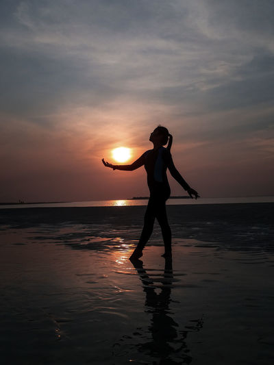 Optical illusion of silhouette woman holding sun while walking against sea during sunset