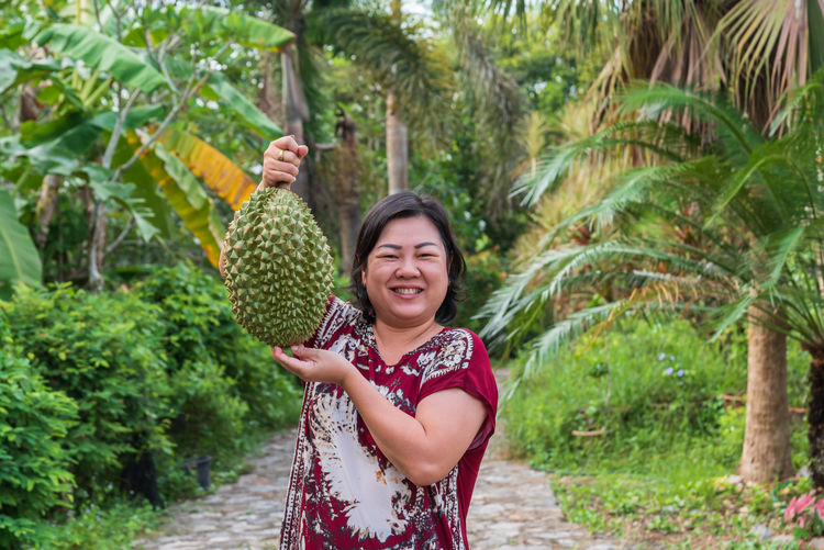 Portrait of happy mature woman holding durian while standing on footpath amidst plants