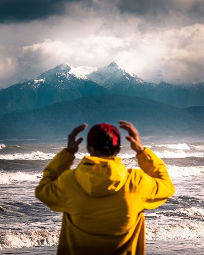 Rear view of young man standing at beach against snowcapped mountains