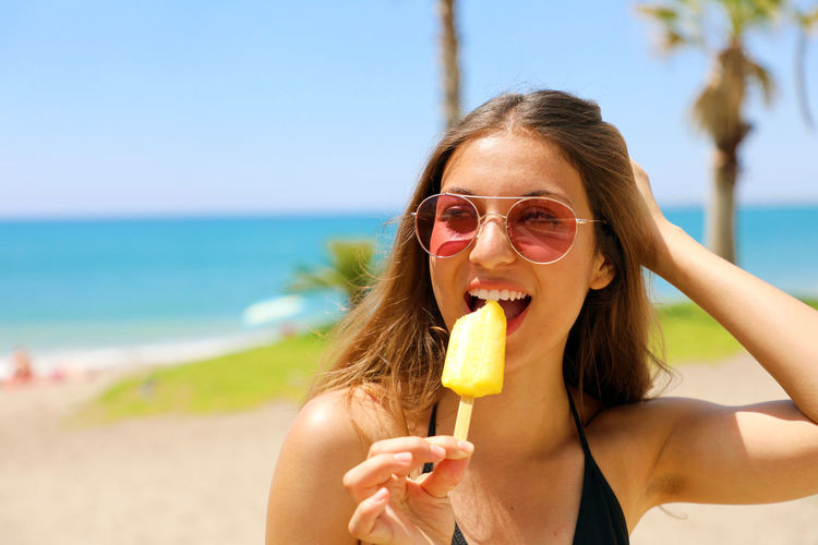 Young woman in sunglasses eating popsicle while standing at beach during sunny day