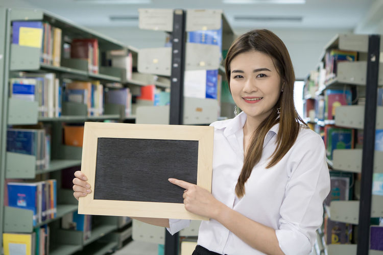 Portrait of smiling young woman pointing at writing slate in library