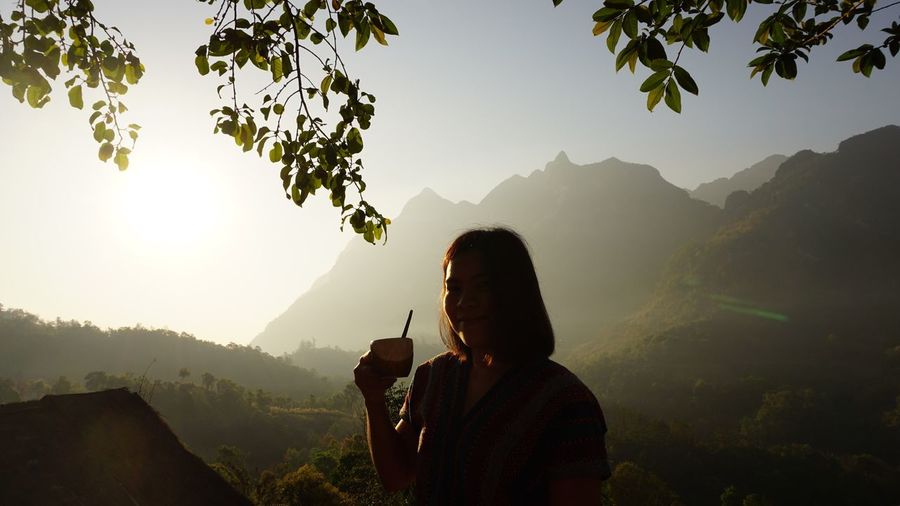 Portrait of woman holding coffee cup by mountains against sky