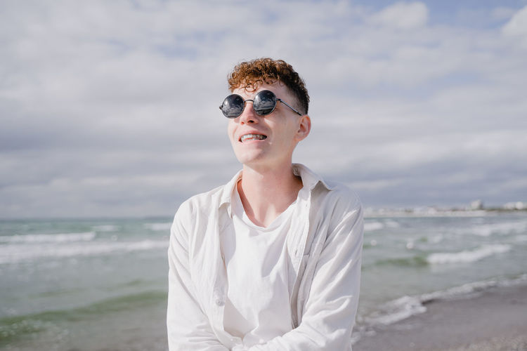 A young guy with braces on his teeth, curly hair and round sunglasses on his face, stands on the sea