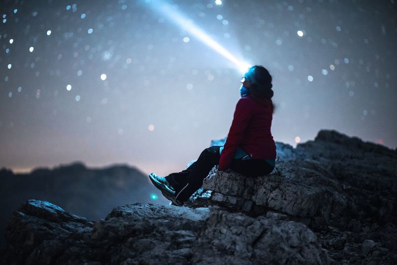Woman wearing headlamp sitting on rock against sky