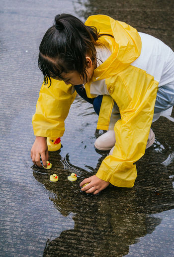Charming ethnic kid in raincoat playing with plastic ducks reflecting in rippled puddle in rainy weather