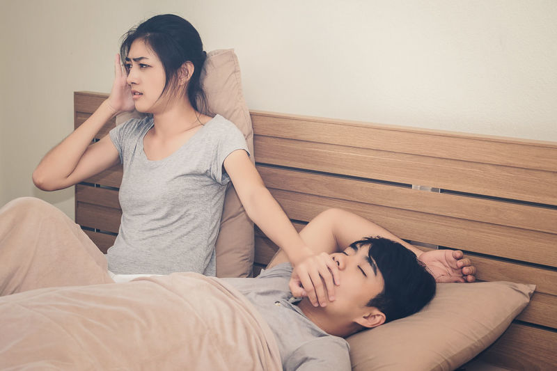 Frustrated woman sitting by man sleeping on bed at home