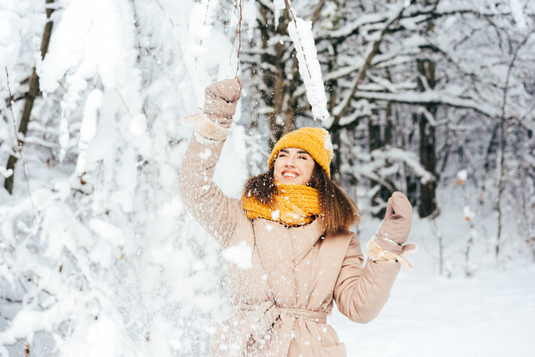 A charming, playful woman in a yellow hat shakes snow from a tree to make it snow.