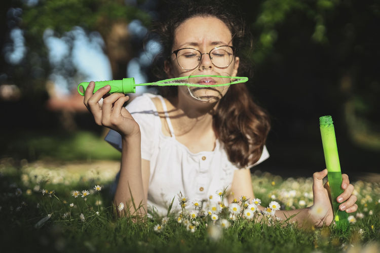 Portrait of a beautiful caucasian teenager girl blowing soap bubbles while lying on a flower meadow