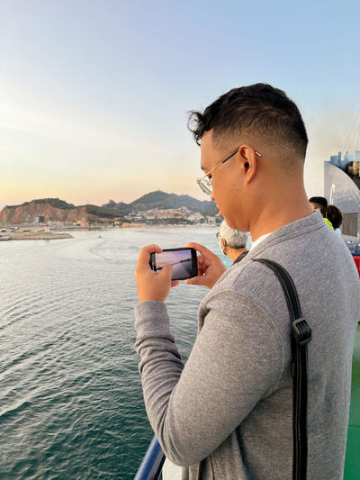 Side view of young man photographing against sky during sunset