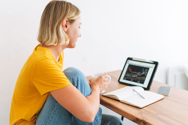 Young woman in yellow t-shirt studies cryptocurrency charts and stock quotes on tablet at home