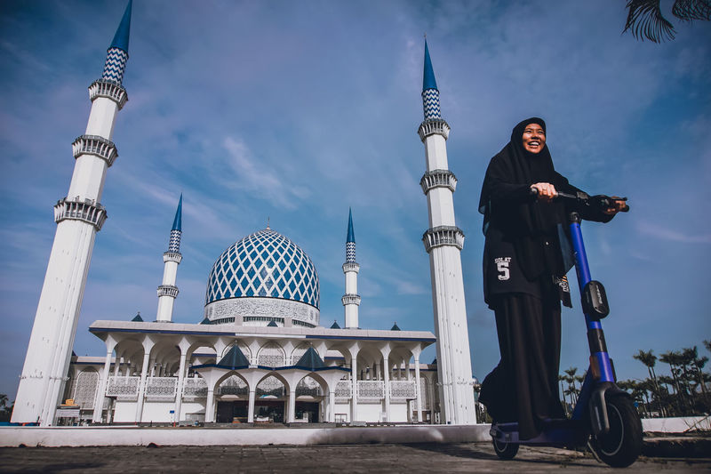 Low angle view of smiling woman riding push scooter against mosque