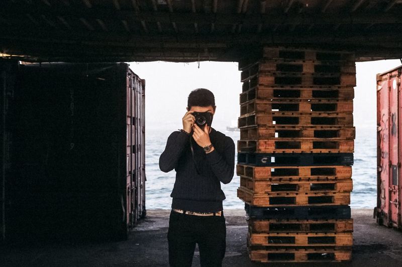 Man photographing through camera while standing against stacked crate