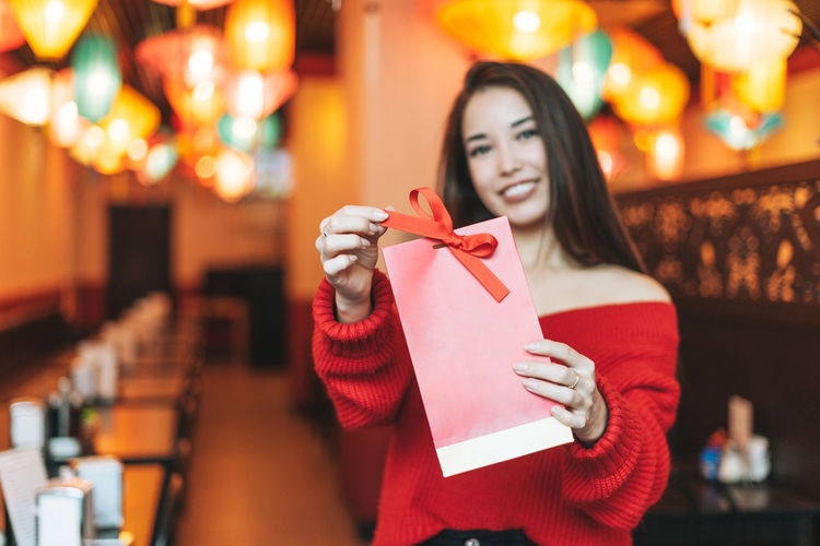 Beautiful young asian woman in red clothes with red gift bag in restaurant celebrating 