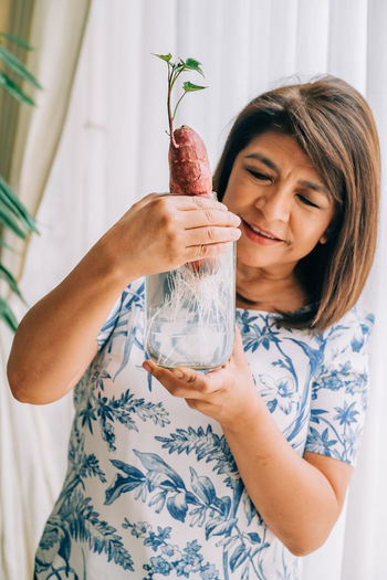 Hispanic woman holding and checking a sweet potato grown in a glass jar filled with water. 