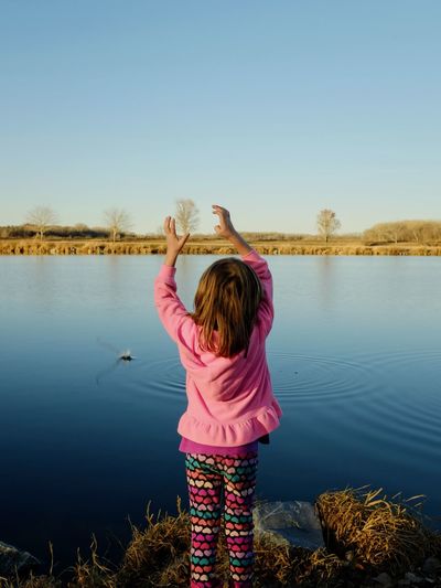 Rear view of girl with arms raised standing at lake against clear sky