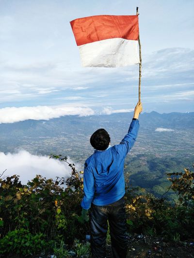 Rear view of man holding indonesian flag while standing on mountain against sky