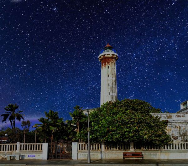 Lighthouse amidst trees and building against sky at night