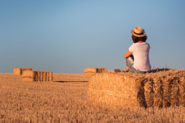 Woman sitting on hay bale against clear sky