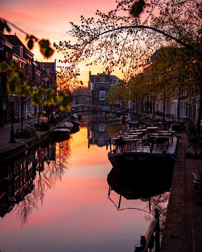 Boats moored in canal by buildings against sky during sunset