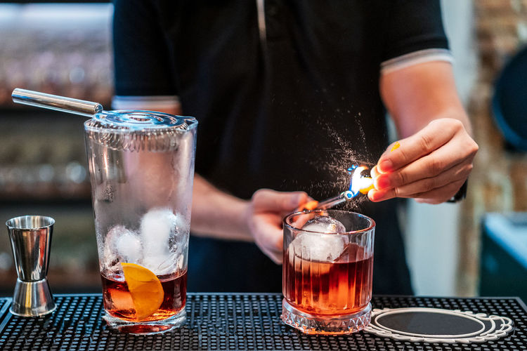 Unrecognizable male bartender preparing negroni cocktail and setting orange slice on fire while standing at counter