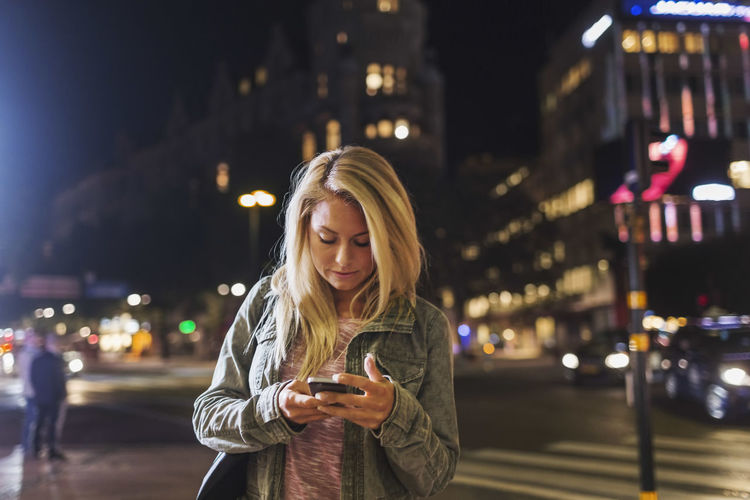 Young woman using phone on street in city at night