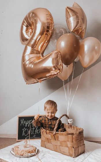 Boy holding balloons while sitting in basket by cake at home