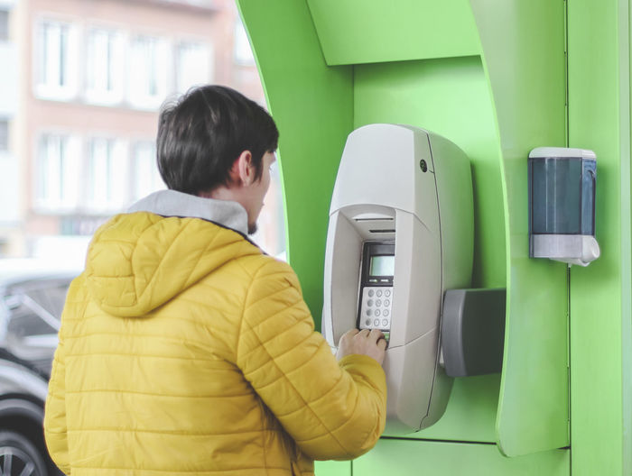 A caucasian guy in a yellow jacket stands near a green atm at a gas station and pays for gasoline