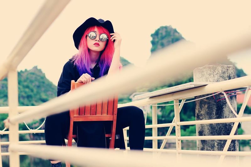 Portrait of young woman wearing sunglasses while sitting on chair by railing