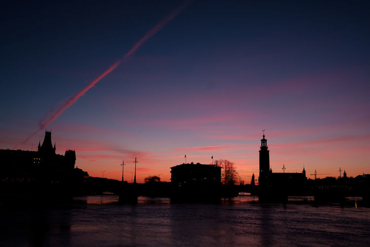 Silhouette buildings by river against sky at sunset