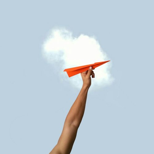 Low angle view of cropped hand holding orange paper rocket against sky