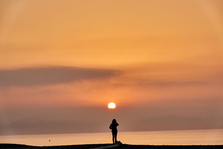 Silhouette person standing by sea against orange sky during sunrise