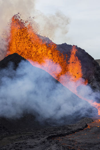 Splashes of hot orange lava erupting from volcanic mountain peak surrounded with smoke in iceland