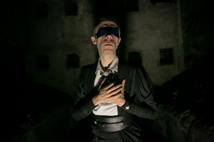 Man with blindfold touching chest while standing against wall at night