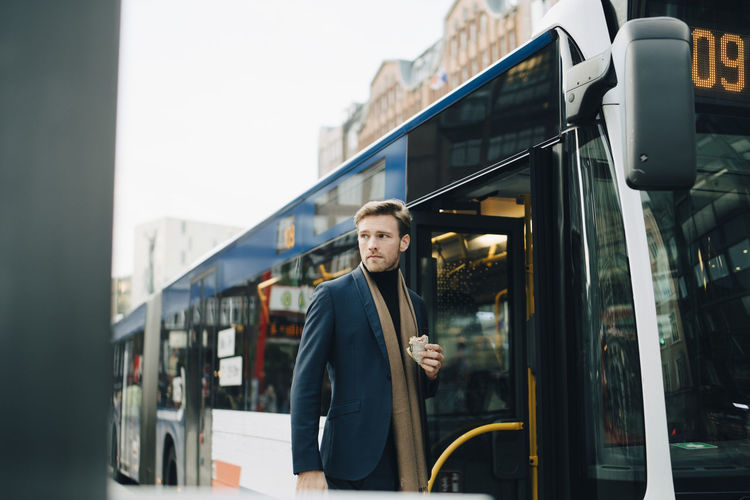 Businessman with wrap sandwich looking away while standing against bus in city