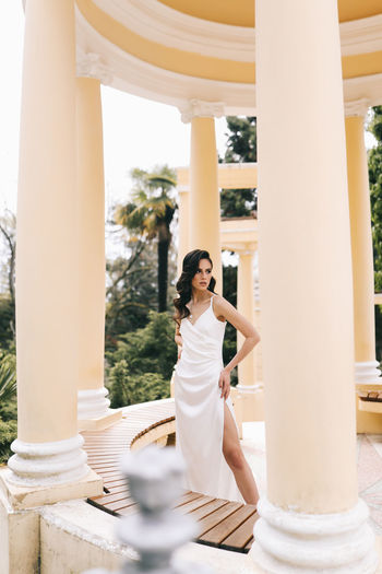 A beautiful brunette lady in an elegant wedding dress poses among the columns in the old city park