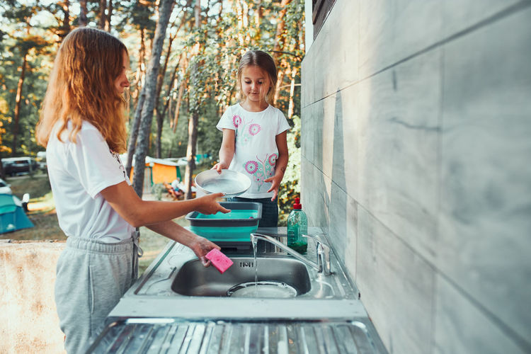 Teenager girl washing up the dishes pots and plates with help her younger sister