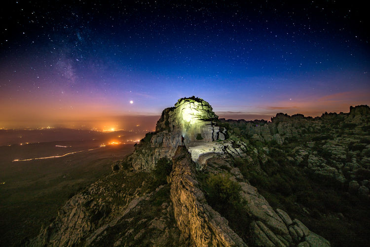 Hiker with illuminated flashlight standing on rock formation against sky at night