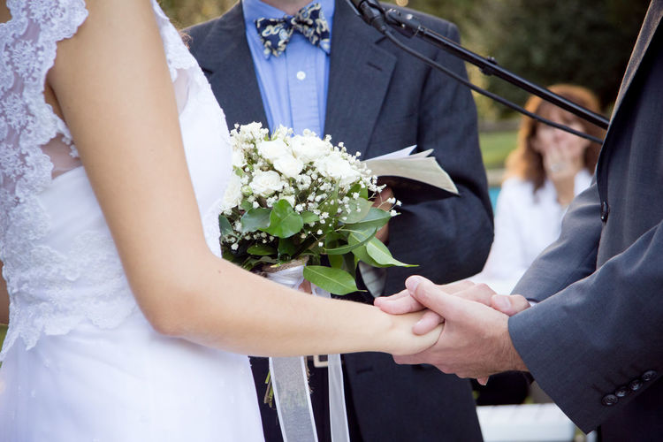 Midsection of bride and bridegroom holding hands by priest during wedding ceremony