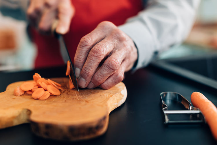 Close up on the wrinkled hands of an elderly man who with a knife and cutting board cuts carrots