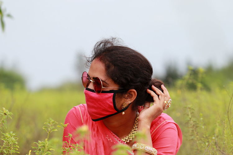 Young woman wearing mask looking away while sitting on field against sky