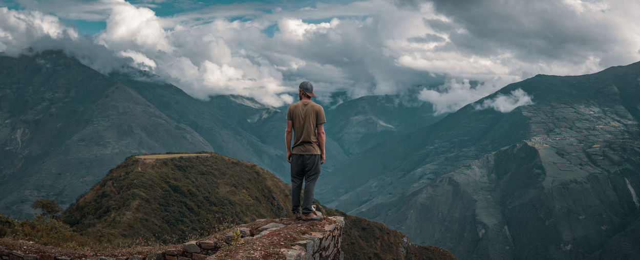 Rear view of young man looking at mountains while standing against cloudy sky