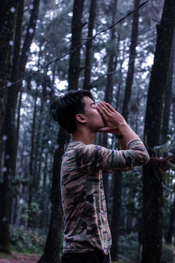 Side view of young man standing against trees in forest