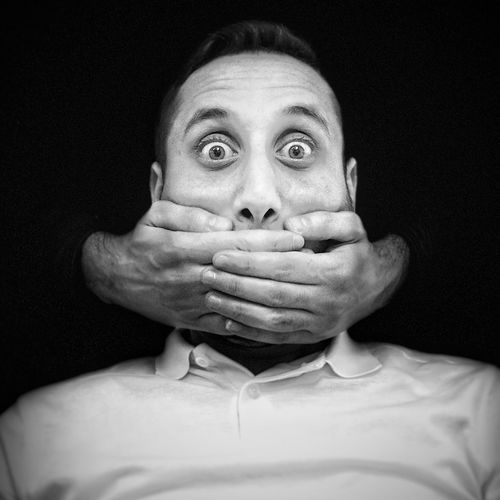 Cropped hands of person covering mouth of man against black background