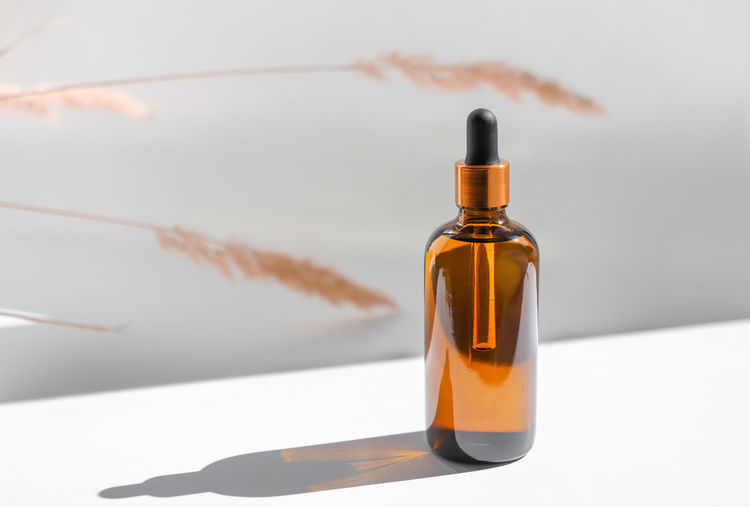 Amber glass bottle with cosmetic body oil on a white background with sunbeams and dry plants. 