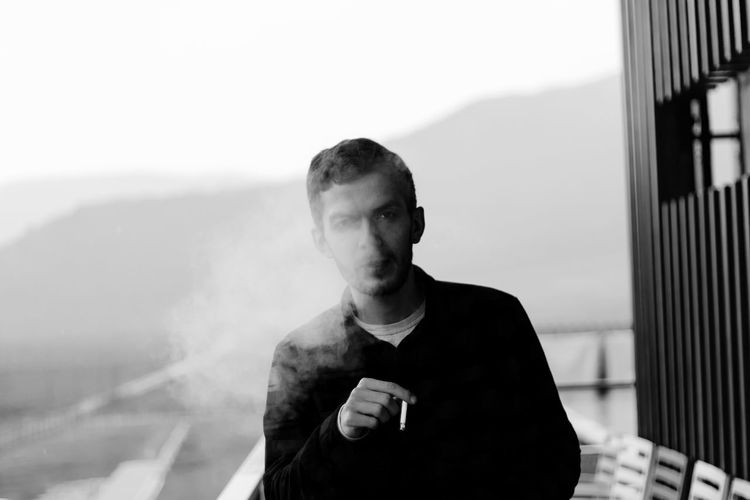 Portrait of handsome young man smoking cigarette against clear sky