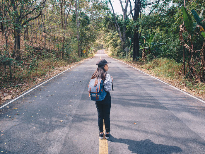 Woman hiking with backpack walking on roadway amid greenery trees.